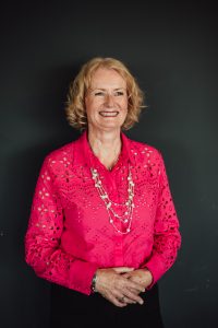 Headshot portrait of a lady wearing pink blouse against a blue wall