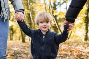 Close up portrait of young boy holding parents hands either side of him in an autumn woodland