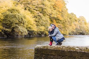 Mum crouching on a stone jetty by a woodland river with young son in autumn