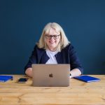 Blond haired female wearing a white top, navy cardy and glasses sat at a wooden desk working on a laptop with a navy blue wall behind her.