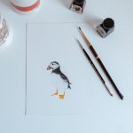 Top down view of a white desk with artists drawing of a penguin with paint and brushes for framing.