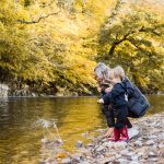 Sister throwing stones into a woodland river in autumn with much younger toddler brother.