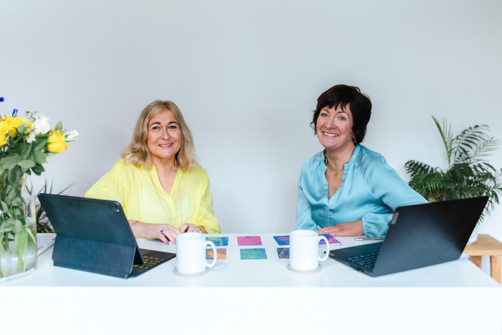 Studio brand shoot with two business women. Both sat at a white desk with white walls behind them.