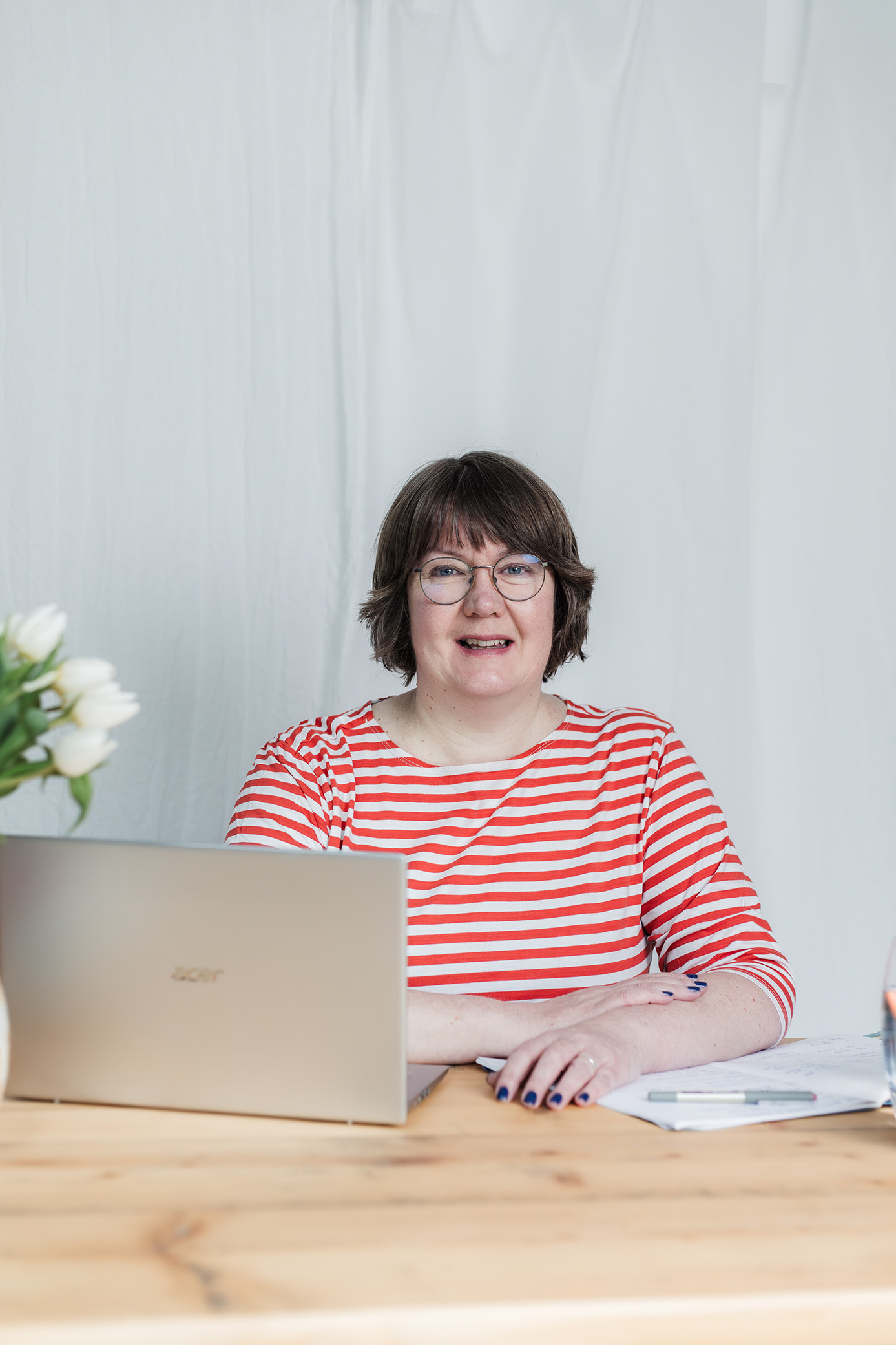 Portrait of a business woman sat at her desk with laptop and white wall behind her.